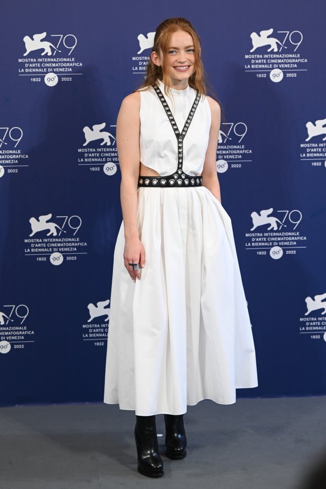 Foto do look da atriz Sadie Sink no Festival de Veneza. Ela usa um vestido branco longo com detalhe de cinto preto e bota de salto alto preta. Ela olha para a câmera e sorri levemente.