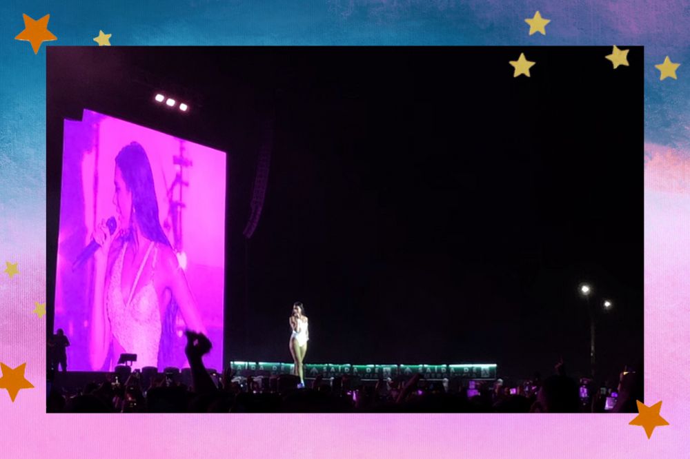 Dua Lipa segurando microfone em palco de show; ao fundo, é possível ver a cantora no telão em uma luz rosa; a margem é uma textura nas cores azul, roxo, rosa, lilás e branco com estrelas amarelas e laranjas decorando a imagem