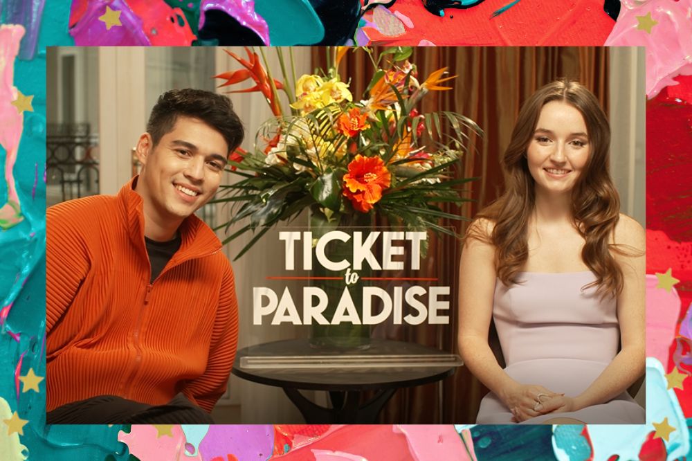 Maxime Bouttier e Kaitlyn Dever, em divulgação para o filme Ingresso Para o Paraíso; eles estão sentados e sorrindo em com o nome do filme em inglês (Ticket To Paradise) está entre eles com um vaso de flores amarelas e laranjas com folhas verdes está no fundo; a margem é uma textura nas cores roxo, verde, vermelho, lilás, rosa, azul e preto com estrelas amarelas como decoração