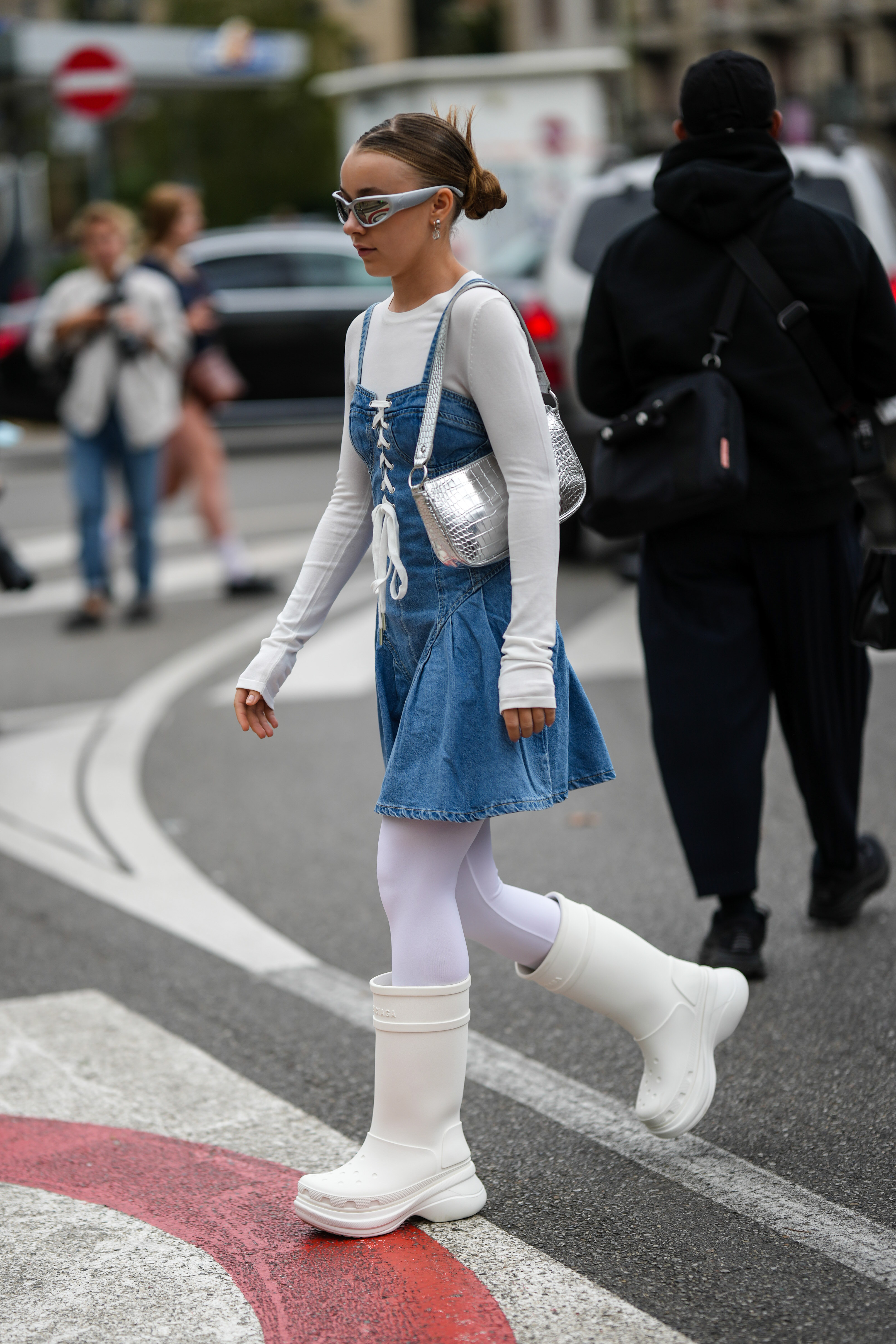 Garota usando vestido jeans por cima de blusa branca, meia-calça branca e galocha branca com óculos futurista e bolsa metalizada no street style da semana de moda de Milão