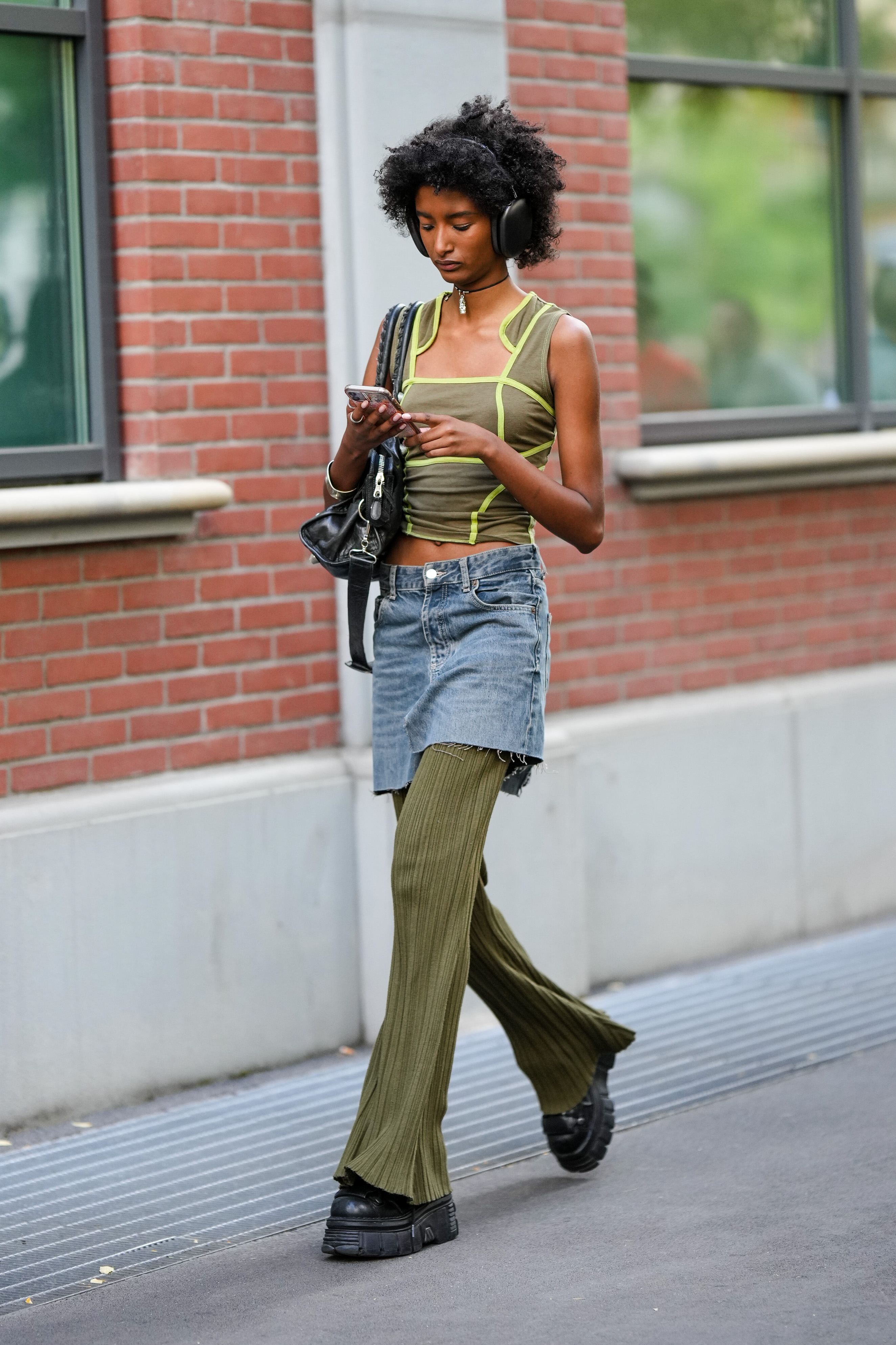 Garota usando top verde, saia jeans por cima de calça flare verde, tênis preto, bolsa preta pendurada em um dos ombros e headphone preto enquanto olha para o celular e anda na rua na semana de moda de Milão