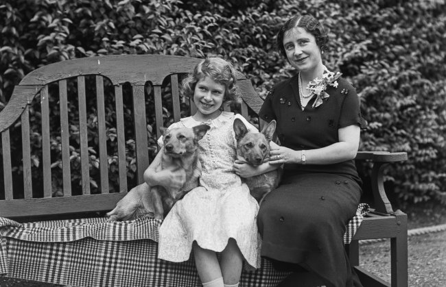 Foto da Rainha Elizabeth II ainda criança, segurança seus primeiros corgis, Dookie e Jane. A imagem é em preto e branco, e a rainha está sentada em um banco ao lado da mãe