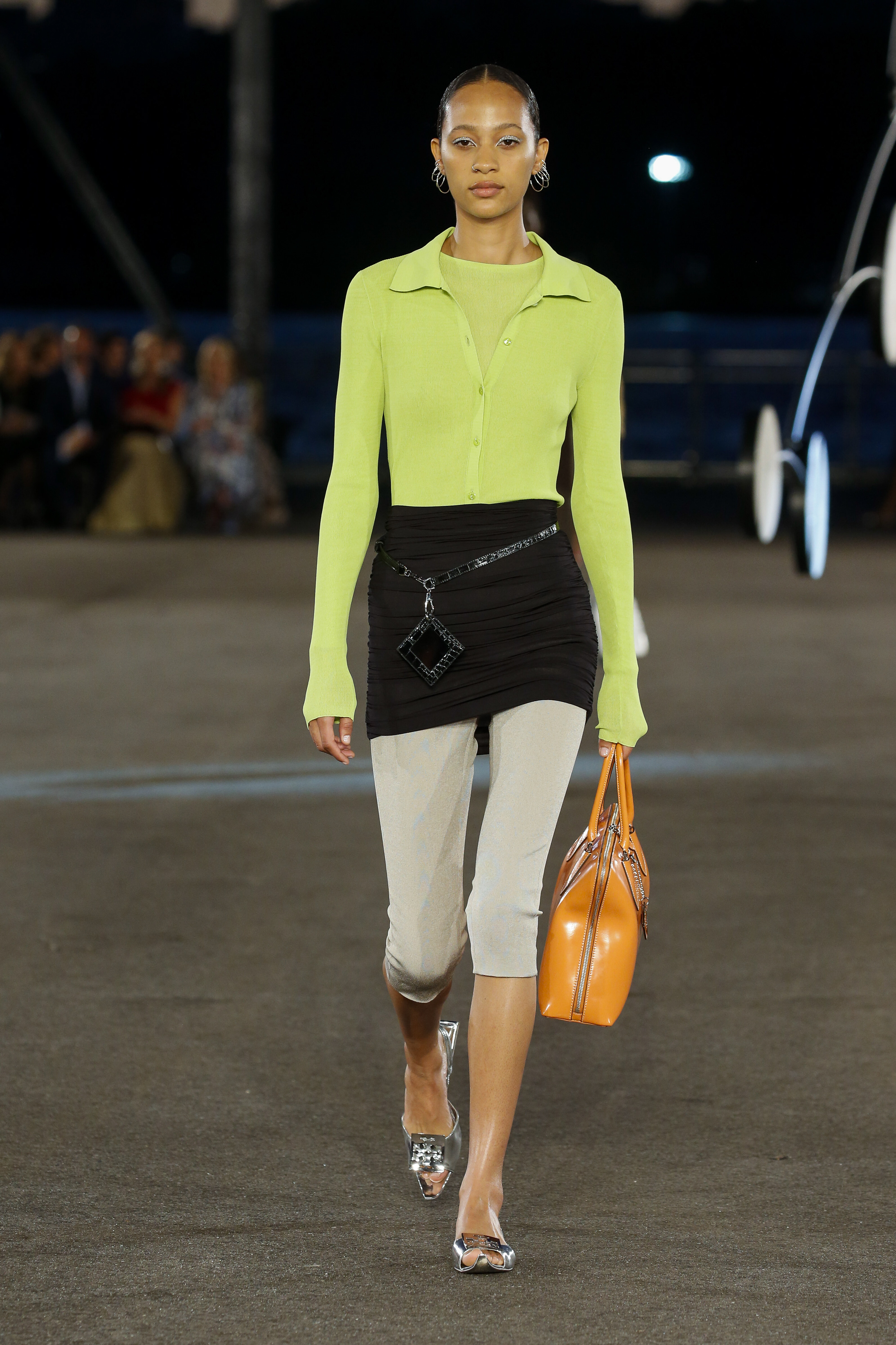Desfile de primavera-verão 2023 da Tory Burch na semana de moda de Nova York. Modelo usando blusa de manga comprida com decote polo em verde neon, saia preta por cima de legging cropped off white e segurando bolsa laranja