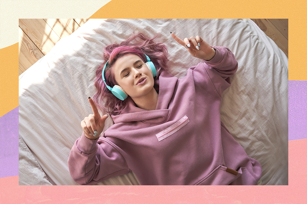 Garota de cabelo rosa ouvindo música com fones de ouvido deitada na cama com os olhos fechados enquanto canta e com as mãos para cima. Montagem em fundo amarelo, lilás e rosa