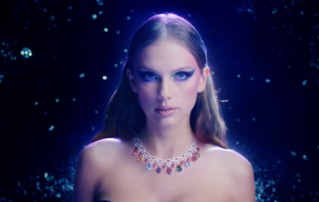 Taylor Swift no clipe de Bejewled; ela está com expressão séria olhando para frente com um colar de pedras coloridas e brilhantes; o fundo é azul escuro com pontos brilhantes se destacando
