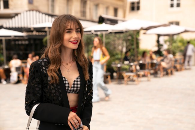 Lily Collins em Emily em Paris; ela está na rua, durante o dia, parada e sorrindo