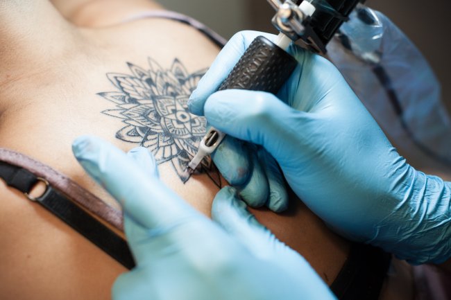 Mão segurando um equipamento de tatuagem e fazendo uma mandala nas costas de uma jovem mulher