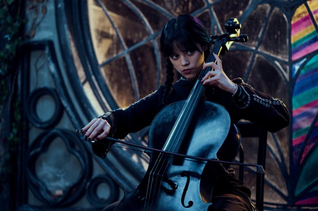 Jenna Ortega como Wandinha Addams em série da Netflix; ela está tocando um violoncelo com uma expressão séria e vestindo roupas pretas