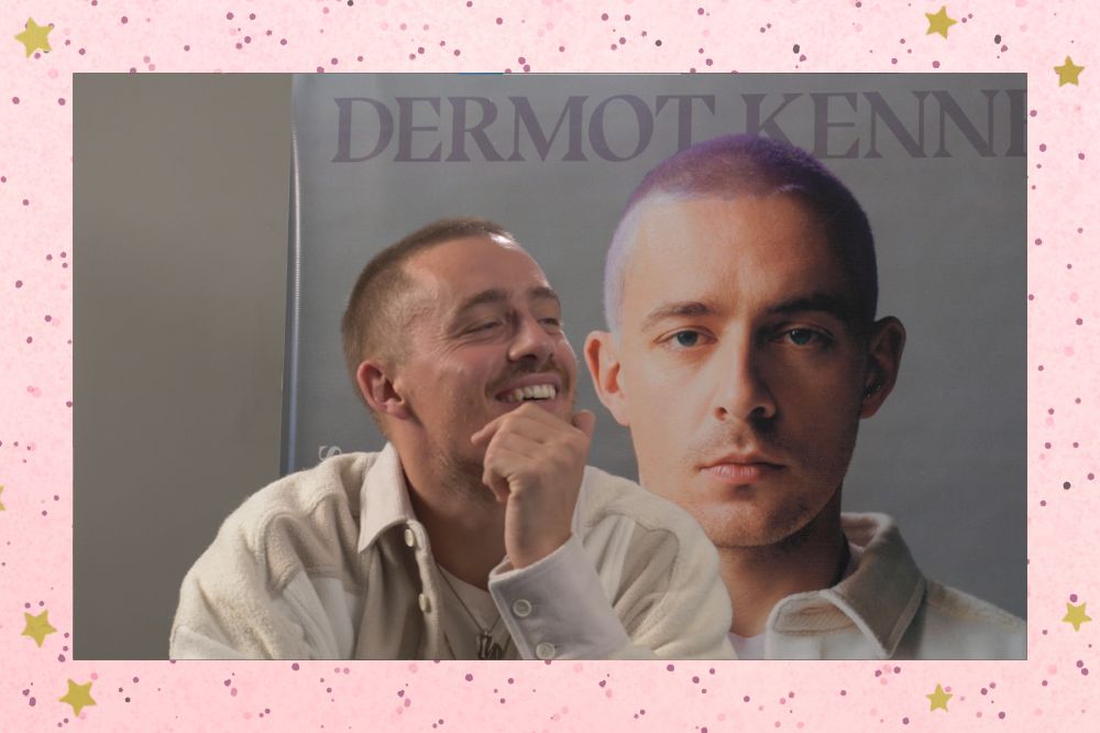 Dermot Kennedy sorrindo com a mão no rosto em entrevista à CAPRICHO; ao fundo é possível ver um banner com a foto do cantor em um fundo cinza na capa do disco SONDER e o nome dele em roxo na parte superior; a margem é uma textura rosa com bolinhas; estrelas amarelas decoram a imagem