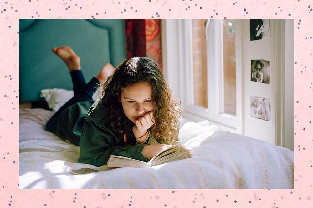 Garota lendo livro no quarto deitada na cama de bruços com os pés para cima
