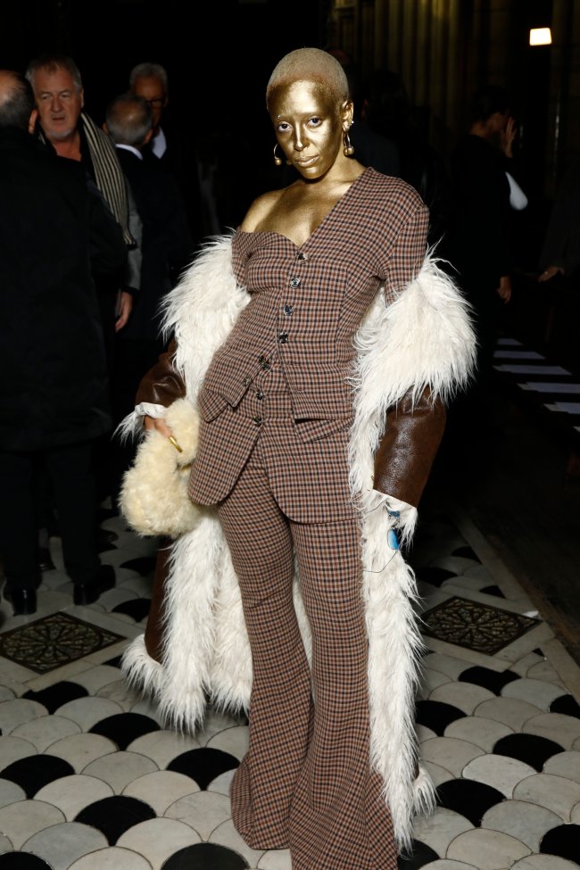 Foto da Doja Cat na Semana de Moda de Paris com um conjuntinho xadrez, casaco marrom de couro e rosto pintado de dourado