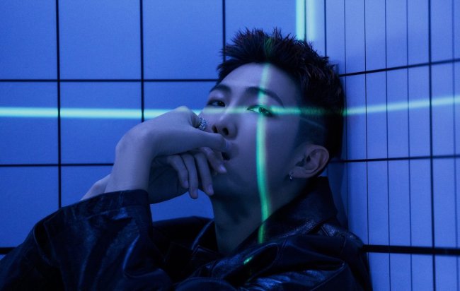 RM posando para foto com a mão no rosto enquanto olha pra câmera e é iluminado por luzes em tons de azul; ele tem expressão séria