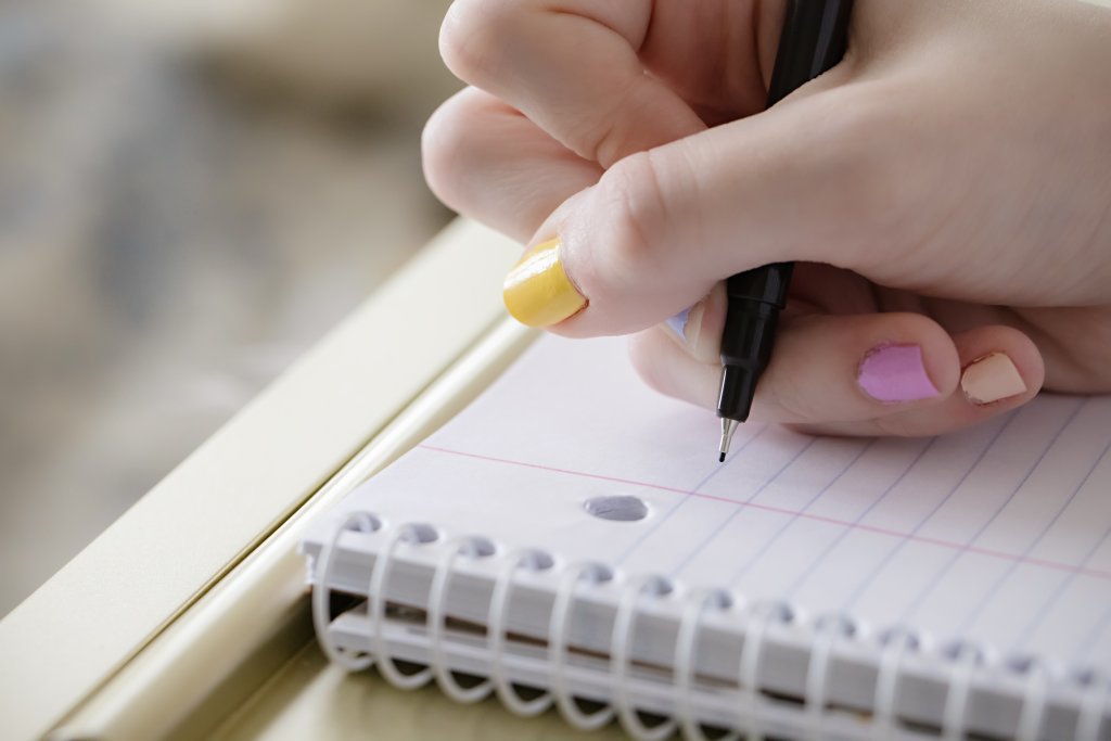 Mão de uma menina escreve em uma folha de caderno. As unhas estão pintadas de amarelo e rosa