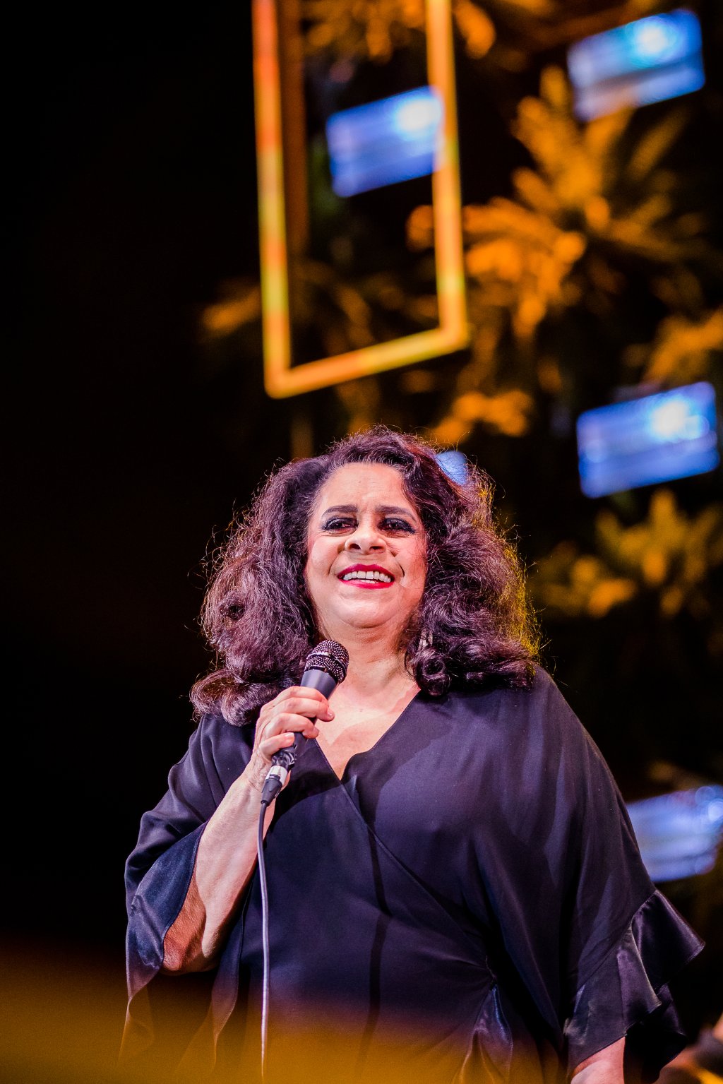 Gal Costa sorrindo enquanto se apresenta no palco com microfone na mão