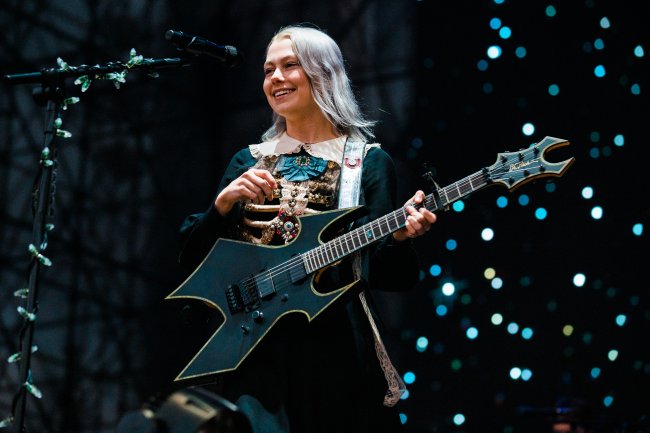 Phoebe Bridgers sorrindo enquanto se apresenta no palco do Primavera Sound em São Paulo; ela observa o público com uma guitarra na frente do corpo enquanto sorri
