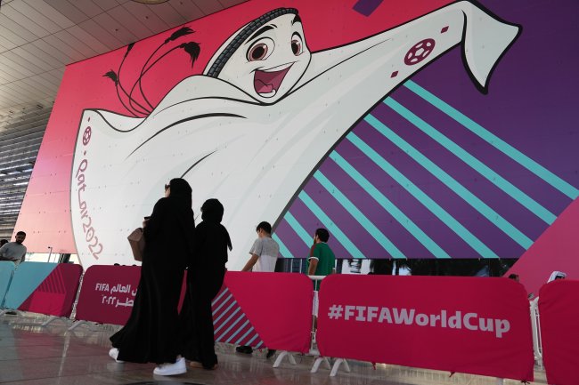Mulheres islâmicas usando hijab passeiam em frente a um cartaz com o mascote da Copa do Mundo 2022