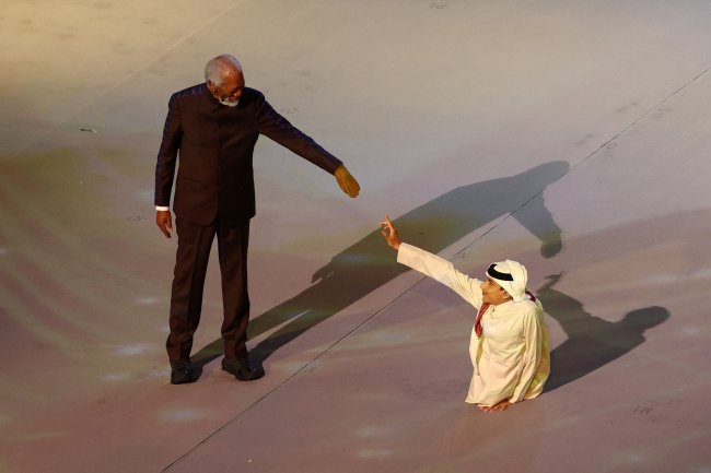 Morgan Freeman e Ghanim Al Muftah se apresentam na cerimônia de abertura da Copa do Mundo 2022, no Catar. Morgan é um homem negro de meia idade e veste um terno. Ghanim é um jovem catari de 20 anos, que não possui as pernas