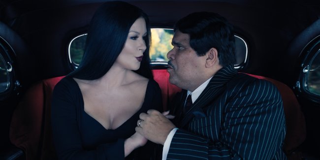 Catherine Zeta-Jones e Luis Guzmán caracterizados como Morticia e Gomez da família Addams; eles estão sentados dentro de um carro de mãos dadas enquanto se olham