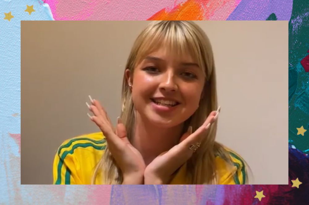 Joalin sorrindo com as mãos próximas ao rosto; ela usa a camiseta do Brasil; a margem é uma textura nas cores rosa, roxo, laranja, lilás e verde; estrelas amarelas decoram a imagem