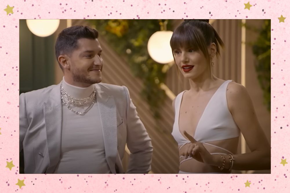 Klebber Toledo e Camila Queiroz em Casamento às Cegas Brasil; ele está olhando para ela e sorrindo levemente enquanto ela aponta com um dos dedos e fala com participantes do reality; a margem é uma textura rosa com bolinhas