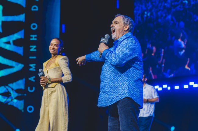 Jon Landau sorrindo e apontando para Zoë Saldaña, que sorri enquanto segura microfone no palco da CCXP22