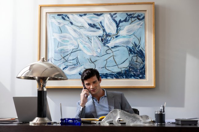 Paul Forman como Nicolas De Leon em cena de Emily em Paris; ele está em um escritório com o celular no ouvido sorrindo levemente; ao fundo um quadro azul, branco e preto decora a parede