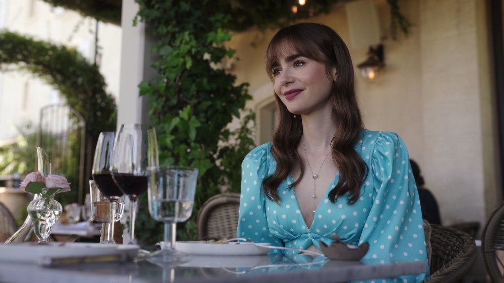 Lily Collins em cena de Emily in Paris; ela está sentada sorrindo levemente enquanto usa uma roupa azul com bolinhas brancas; copos e pratos estão na mesa que está na frente da personagem