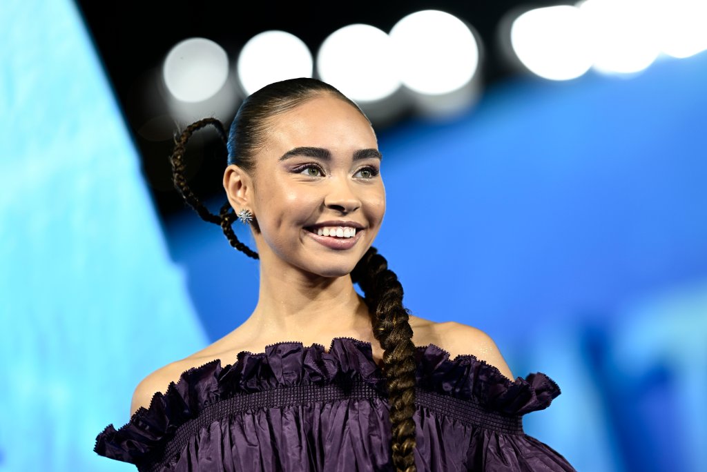 Bailey Bass na premiere de Avatar: O Caminho da Água; ela está usando um vestido roxo e sorrindo; atrás dela, está um fundo azul com luzes