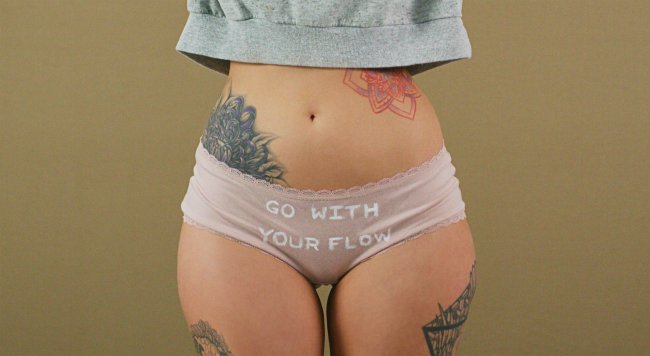 Garota branca e tatuada vestindo uma calcinha rosinha claro com a frase 