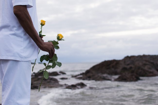 Homem negro vestido de branco parado em frente ao mar, levando na mão rosas amarelas