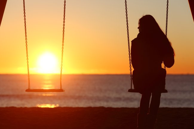 Mulher sentada em uma balanço na praia, durante o pôr do sol. Ele está olhando para o lado, para um balanço vazio, como se sentisse falta de alguém