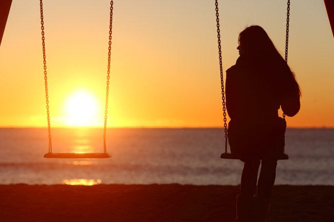 Quais são os tipos de saudade que existem e como lidar com todos eles Mulher sentada em uma balanço na praia, durante o pôr do sol. Ele está olhando para o lado, para um balanço vazio, como se sentisse falta de alguém