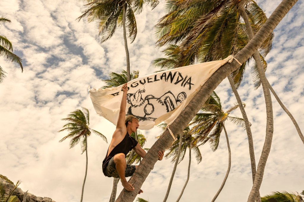 Rudy Pankow como JJ em Outer Banks; ele está rindo e gritando em cima de uma árvore em um dia ensolarado enquanto pendura uma bandeira branca com a palavra "Poguelandia" escrita em preto com um desenho