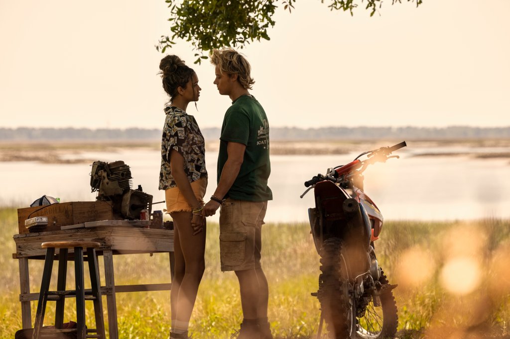 Madison Bailey como Kiara e Rudy Pankow como JJ na 3ª temporada de Outer Banks; eles estão em pé e próximos um do outro em uma área aberta; JJ está com a mão próxima da de Kiara enquanto eles se olham