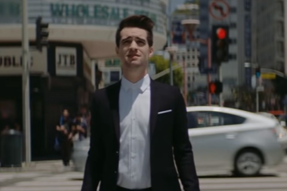 Brendon Urie do Panic! At The Disco em clipe de High Hopes; ele está parado na rua em um dia ensolarado olhando para cima com expressão confusa; ele usa um terno preto com camisa branca