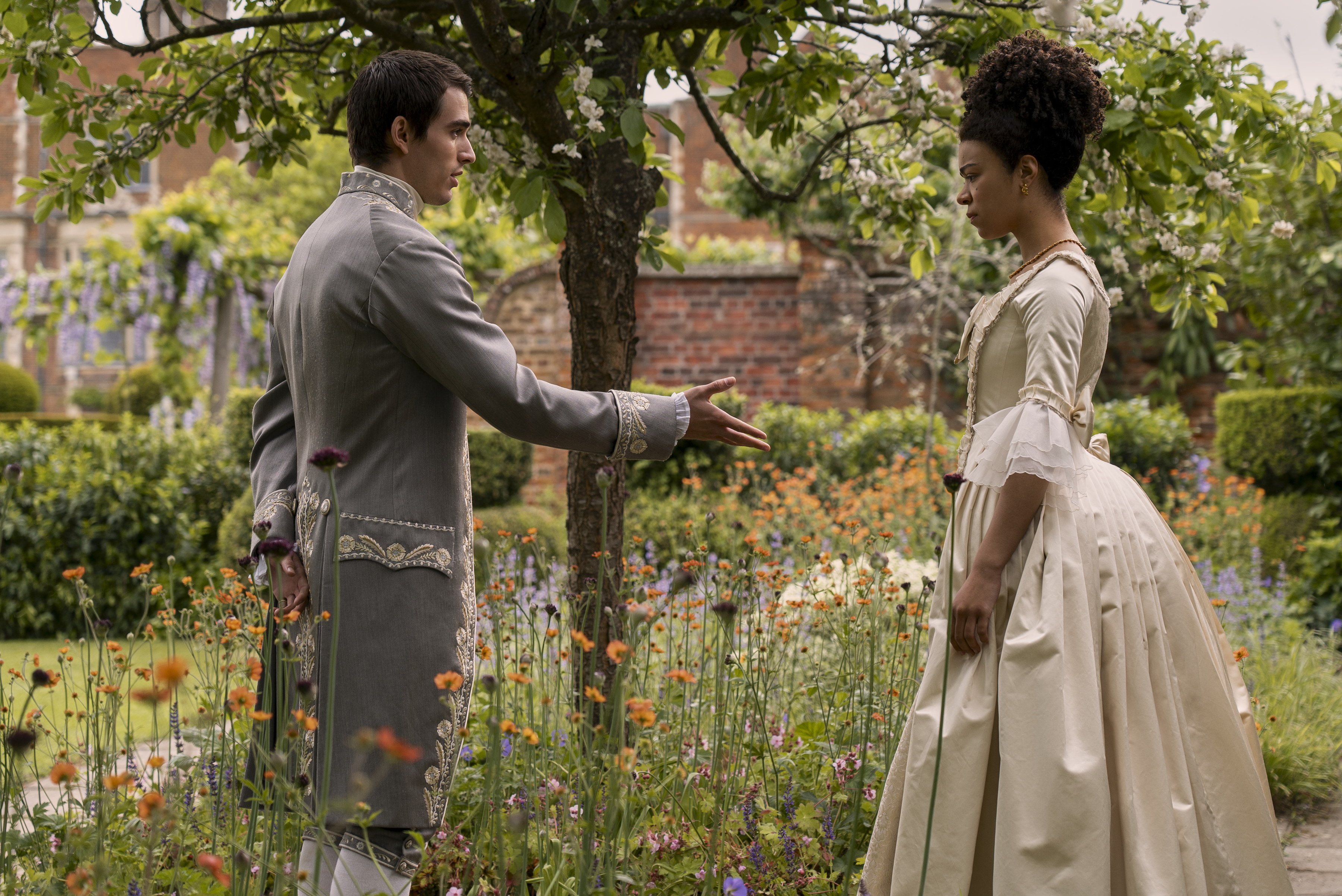 Corey Mylchreest como rei George jovem e India Amarteifio como rainha Charlotte jovem; os dois estão se encarando enquanto em pé em um jardim durante o dia enquanto ele estende a mão para ela