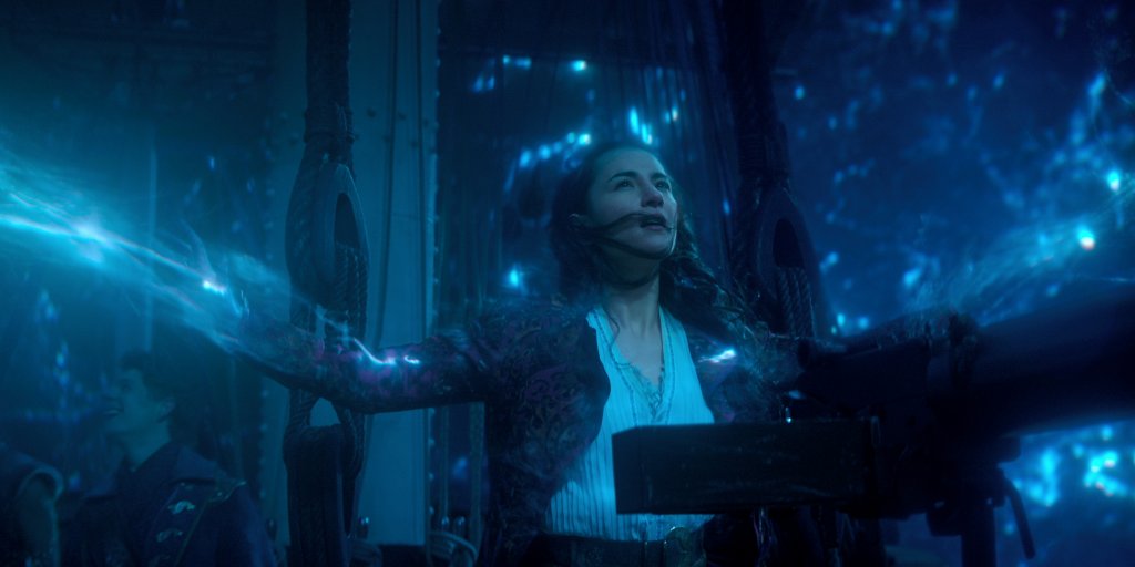 Jessie Mei Li como Alina Starkov em Sombra e Ossos; ela está com os braços abertos enquanto usa seu poder que aparece em uma luz azulada na imagem