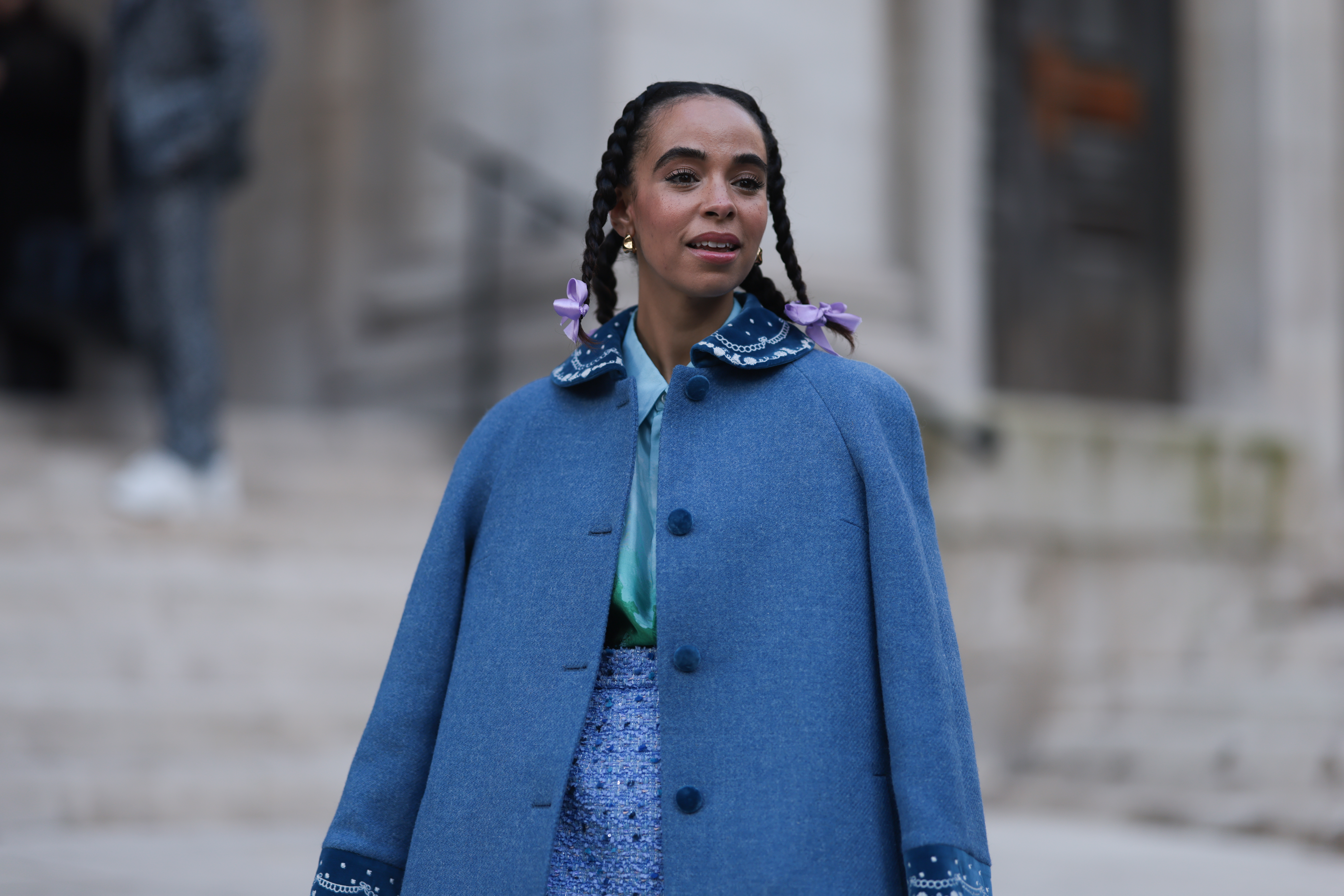 Mulher usando capa azul e cabelo preso em tranças com laços nas pontas no street style da semana de moda de Londres