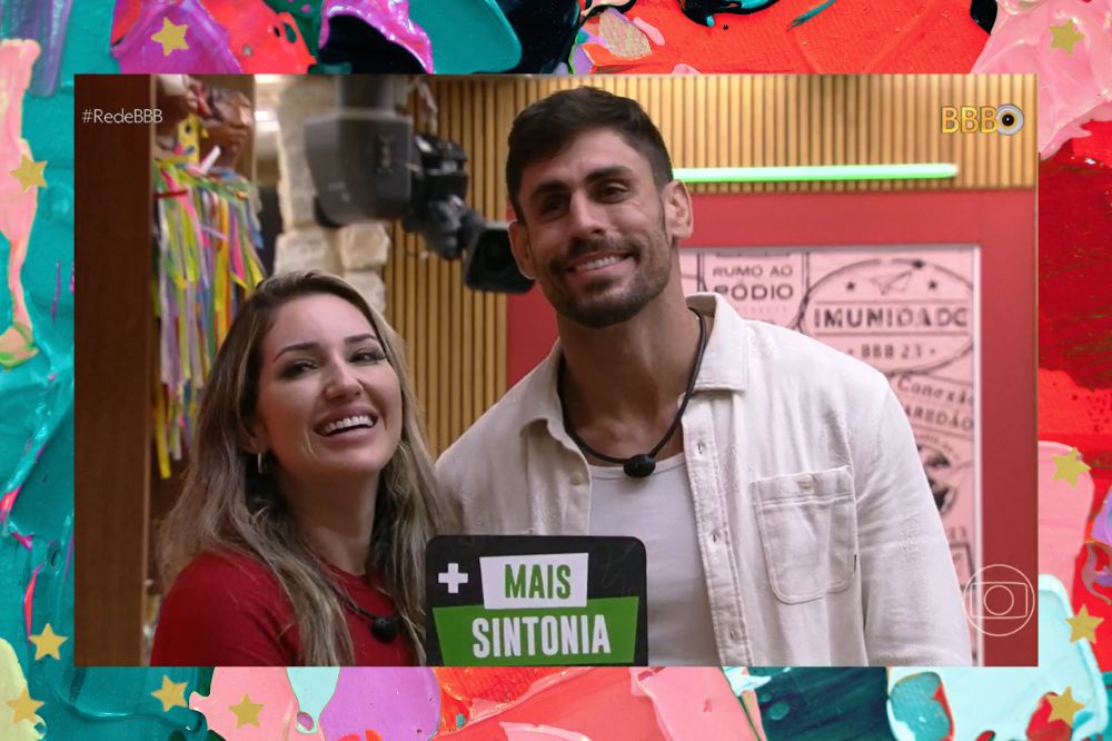 Amanda e Cara de Sapato sorrindo enquanto posam com uma plaquinha dizendo "+Mais Sintonia" na sala do BBB23; a margem é uma textura nas cores verde, roxo, vermelho, lilás, rosa, azul e preto; estrelas amarelas decoram a imagem