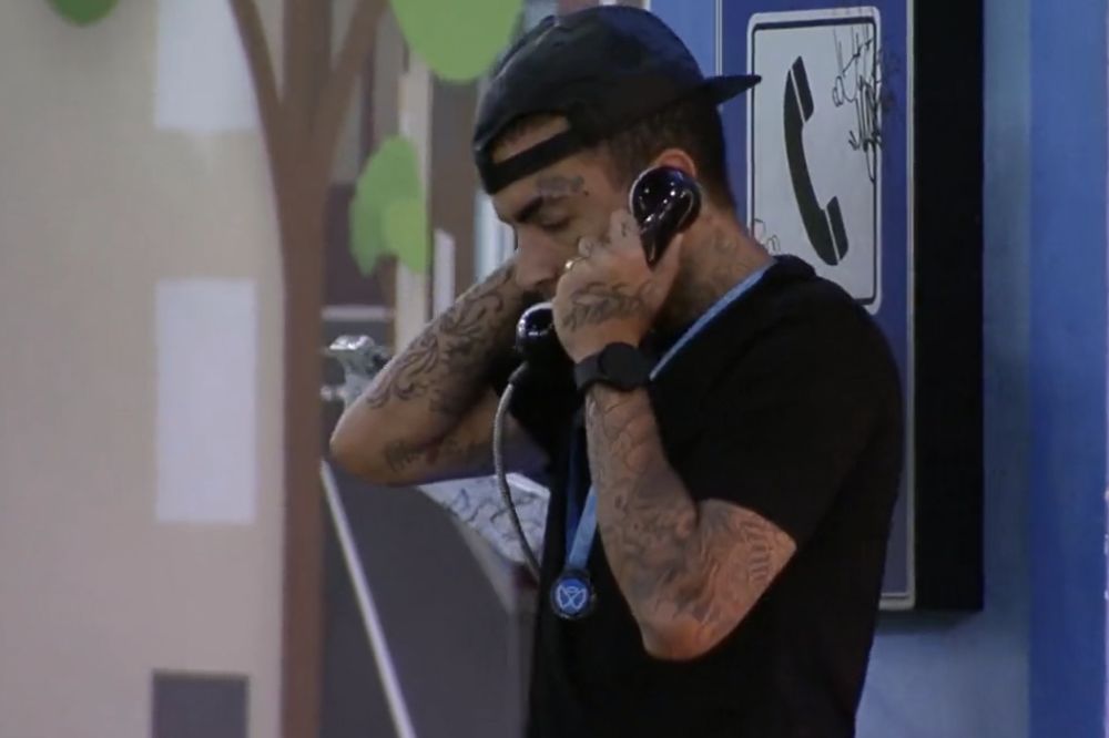 Guimê atende ao Big Fone no BBB23; ele está na área externa segurando o telefone enquanto usa um boné e blusa preta