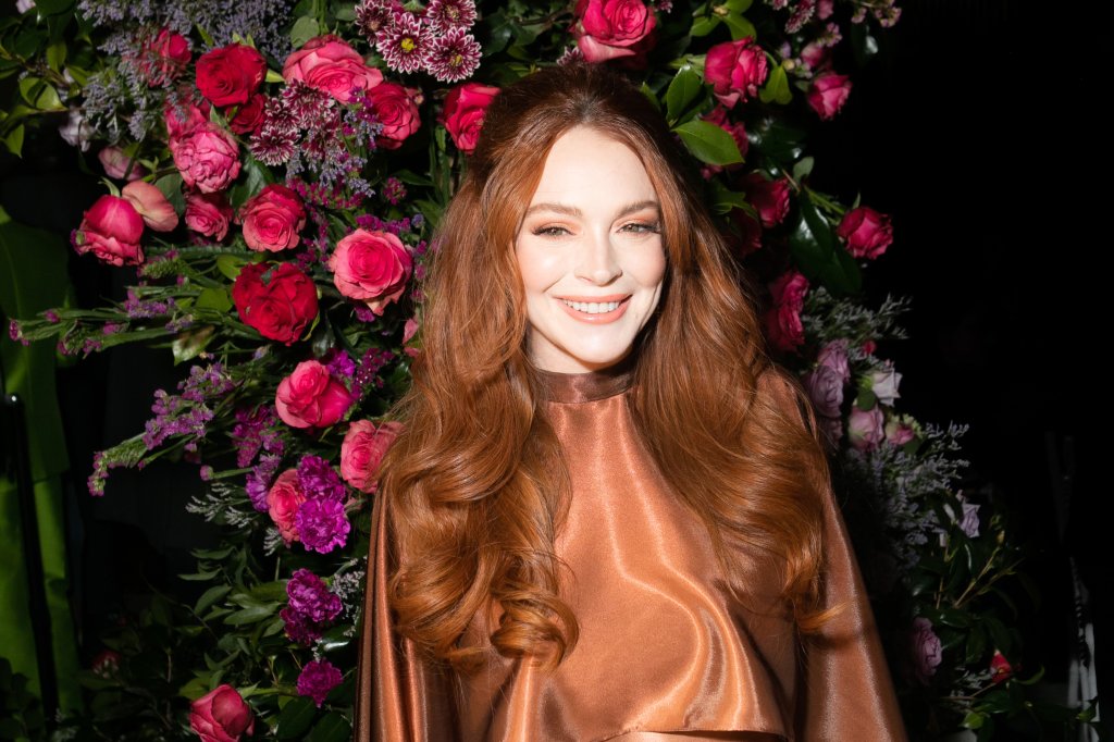 Lindsay Lohan sorrindo para foto com fundo de flores nas cores rosa, vermelho e roxo com folhas verdes