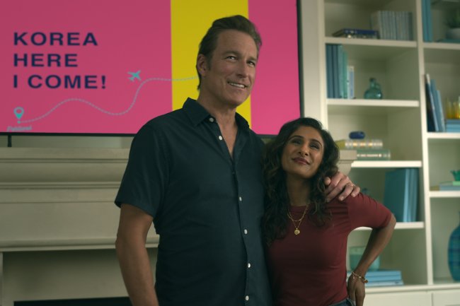 John Corbett como Dan Covey e Sarayu Blue como Trina Rothschild em XO, Kitty; eles estão parados e abraçados enquanto sorriem na sala de uma casa