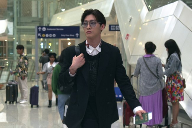 Sang Heon Lee como Min Ho em XO, Kitty; ele está com expressão séria andando por um aeroporto com uma mala