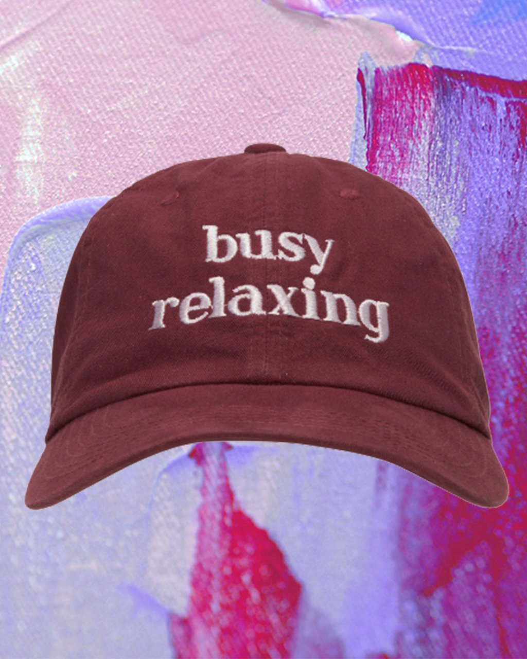 Montagem em fundo lilás e roxo com foto de boné bordô escrito "busy relaxing"