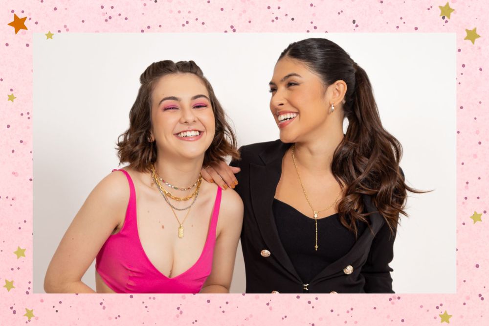 Klara Castanho e Fernanda Concon posando em um fundo branco e sorrindo; Klara Usa um conjunto rosa enquanto Fernanda usa um conjunto preto e está com a mão apoiada no ombro de Klara; a marge é uma textura rosa com bolinhas
