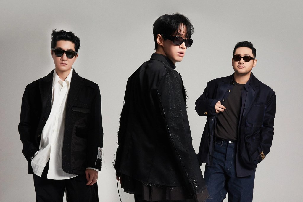 Integrantes do Epik High posando para foto com expressões neutras em um fundo cinza e branco; eles usam roupas pretas e brancas e estão usando também óculos escuros