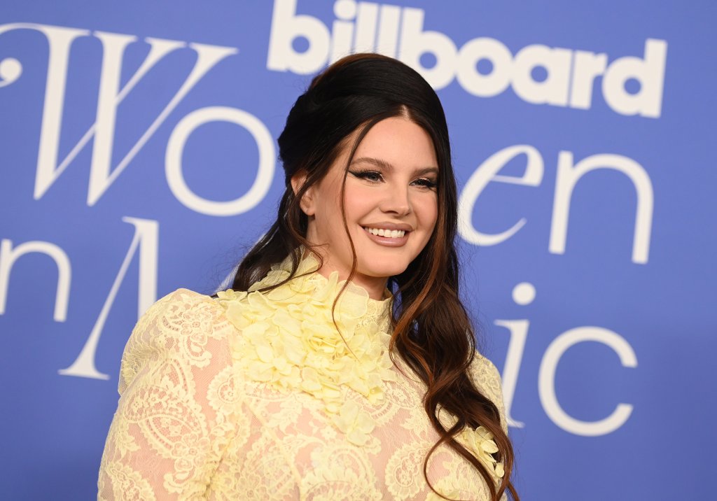 Lana Del Rey no Billboard Women In Music 2023; ela está posando para foto sorrindo com um vestido amarelo com um backdrop com o nome do evento em azul ao fundo