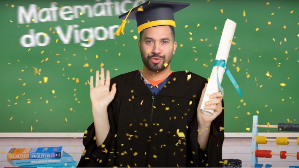 Na imagem aparece Gil do Vigor usando uma beca e um capelo. Em uma das mãos ele segura um diploma. No fundo da imagem aparece uma lousa verde com o escrito "Matemática do Vigor"