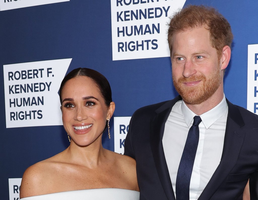 Meghan Markle e príncipe Harry posando para foto sorrindo em um backdrop azul com o nome do evento 2022 Robert F. Kennedy Human Rights Ripple Of Hope Gala em Nova Iorque