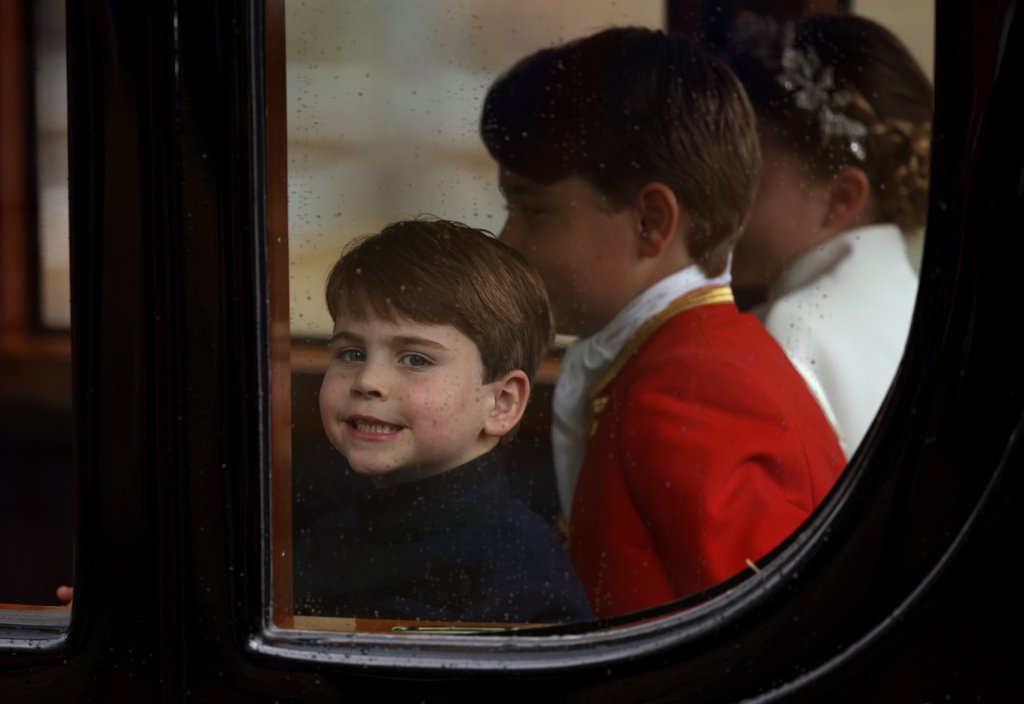 Príncipe Louis, príncipe George e princesa Charlotte no dia da coroação do Rei Charles III; eles estão sentados dentro de uma carruagem com Louis olhando pela janela e sorrindo